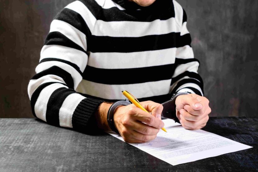 Handcuffed man in a striped shirt filling out a document with a yellow pen.
