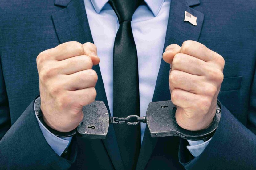Man in a blue business suit wearing metal handcuffs with both fists raised.