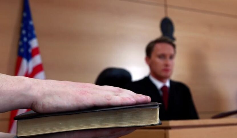Witness placing a hand on the Bible to take an oath before testifying in a courtroom, with a judge seated in the background.